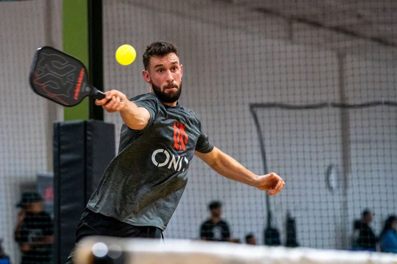 A man playing pickleball indoors, wearing a gray shirt and shorts, is using the Onix Evoke Premier Pro Raw Carbon Pickleball Paddle to hit a yellow ball. In the background, there's a net and blurred figures watching the game.