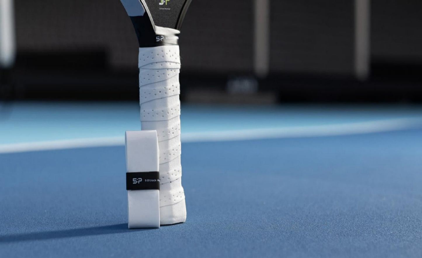 A tennis racket with a white grip stands upright on a blue court. Next to it is a packaged 5 O'clock Pickleball Picklepacks grip tape labeled SP, ideal for enthusiasts who enjoy customizing their paddles. The background is blurred, highlighting the game-day equipment.