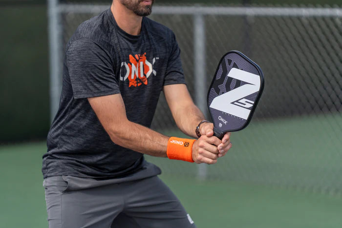 A person confidently stands on a pickleball court, framed by a chain-link fence, holding an Onix Graphite Z5 v3 pickleball paddle while wearing a black shirt with an orange graphic and matching orange wristband.