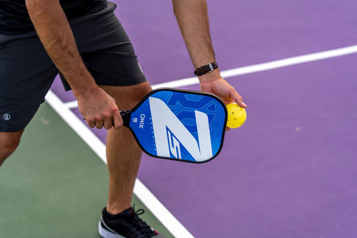 A person wearing black shorts prepares to hit a yellow pickleball with their Onix Graphite Z5 v3 Pickleball Paddle, which features a tennis handle shape, on a court with green and purple sections.