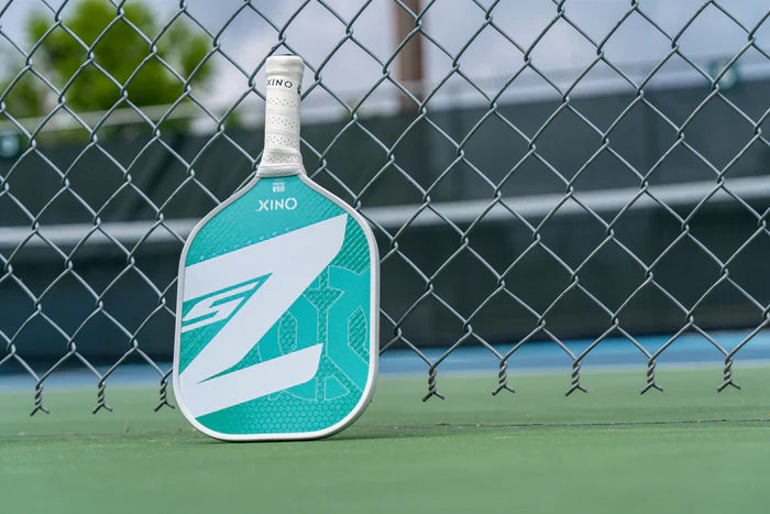 A Graphite Z5 v3 Pickleball Paddle by Onix, showcasing a teal and white design with the word XINO on its tennis handle shape, leans against a chain-link fence. The scene is set on a court with a blurred background, highlighting the sleek look of this paddle.