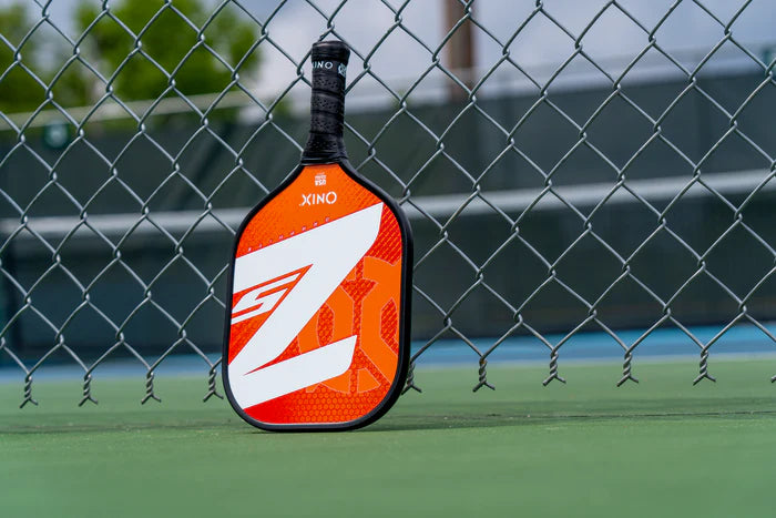 An Onix Graphite Z5 v3 pickleball paddle, featuring a red and black design with a distinctive white Z logo, is positioned upright against a chain-link fence on the tennis court. Its sophisticated design is subtly emphasized as the background, with slightly blurred court lines and a touch of greenery, frames the scene.