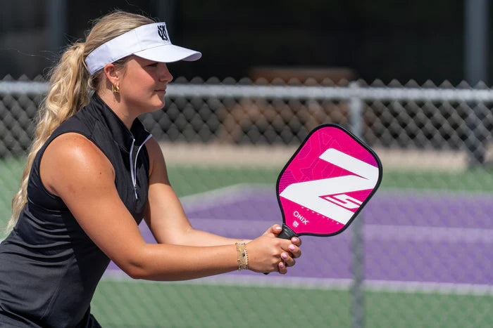 A woman in a visor and sleeveless top grips a pink and white paddle with a tennis handle shape, ready to hit the ball. She's on a pickleball court featuring a purple surface with a chain-link fence in the background, showcasing her trusty Onix Graphite Z5 v3 Pickleball Paddle.