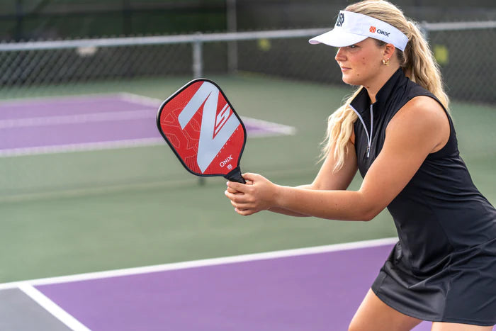 A woman in a visor and black sleeveless dress is ready to play pickleball on the court. She grips her Onix Graphite Z5 v3 Pickleball Paddle, with its striking red and black design, as she stands near the net, prepared to return the ball.