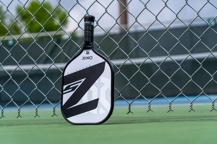 A black and white Graphite Z5 v3 Pickleball Paddle by Onix, featuring a tennis handle shape, rests against a chain-link fence on a green court. The background reveals a cloudy sky and blurred tennis courts.