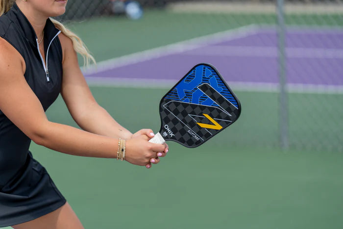 A person dressed in a black outfit is holding an Onix Graphite Z7 v2 Pickleball Paddle on a court. The USA Pickleball approved paddle showcases a sleek black and blue design with striking orange accents. In the background, the net and purple court area can be seen.