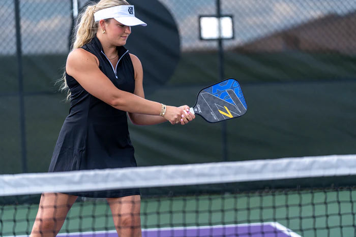 A woman dressed in a black dress and white visor is playing pickleball on an outdoor court, skillfully using the Onix Graphite Z7 v2 Pickleball Paddle, which is USA Pickleball approved. Encircled by a black chain-link fence, her focus and expert paddle technique are highlighted as she prepares to hit the ball.