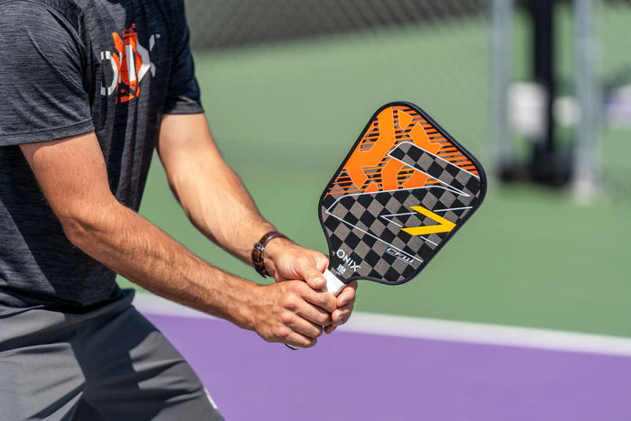 Standing ready behind the net on a purple court, a person wearing a dark shirt and gray shorts grips the Onix Graphite Z7 v2 Pickleball Paddle, known for its geometric orange and black design. The USA Pickleball Approved, lightweight carbon fiber construction ensures swift and agile movements during play.