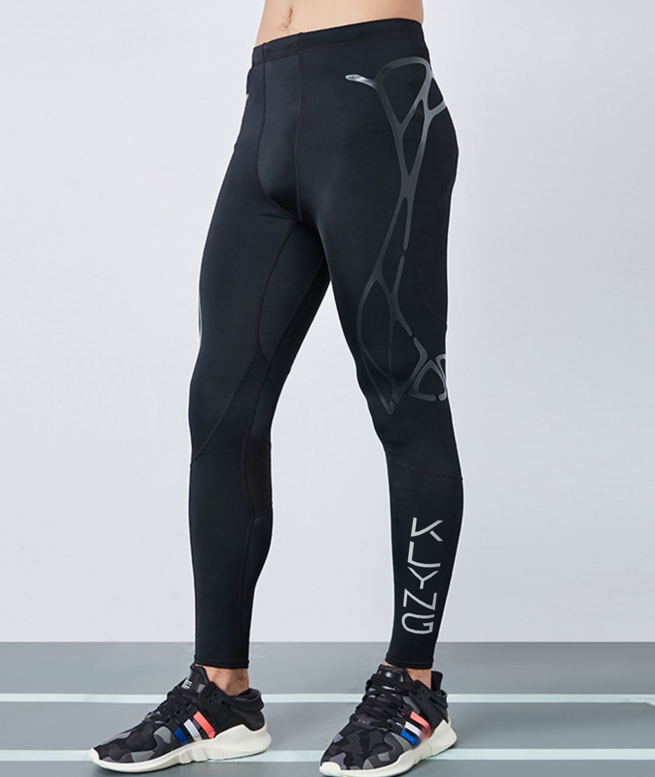 A person wearing black athletic leggings with a geometric design on the left thigh and "Klyng" printed on the right shin, enhanced with Men’s PowerFlex Pickleball Compression Pants for optimal muscle support. Paired with black sneakers featuring colorful stripes against a light gray wall.