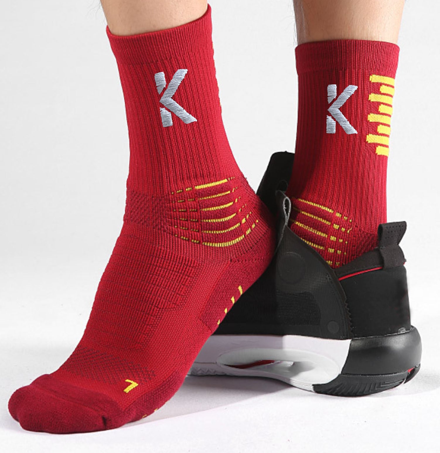 A person is standing in black sneakers with white soles against a plain background, wearing Klyng's PowerStep Achilles Support Compression Socks in red. These unisex socks feature a white "K" logo and yellow accents, making them ideal for a pickleball game.