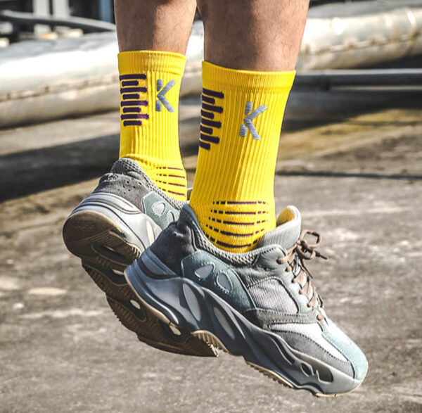 Wearing Klyng's PowerStep Achilles Support Compression Socks in a vivid yellow pattern, a person mid-jump in gray sneakers stands out against the industrial backdrop. The sunny outdoor setting highlights the effectiveness of the vibrant unisex footwear.