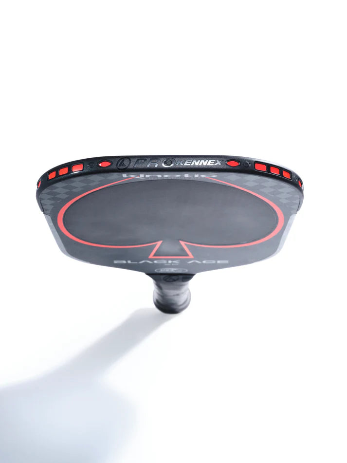 Close-up of an elongated Prokennex Black Ace XF Pickleball Paddle viewed from the top edge, highlighting the logo and design details. The paddle features a black and red color scheme with a textured surface and a thin core, along with a visible handle at the bottom against a white background.