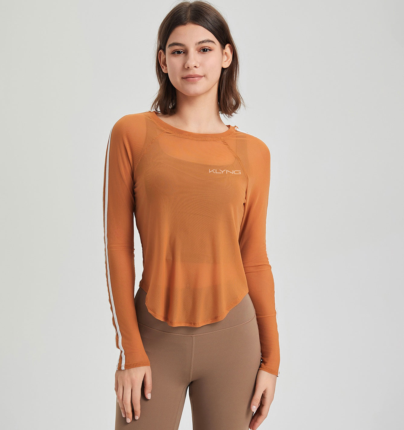 A person wearing a sheer long-sleeve orange SwiftMesh Performance Top by Klyng and brown leggings stands against a plain background, looking directly at the camera.