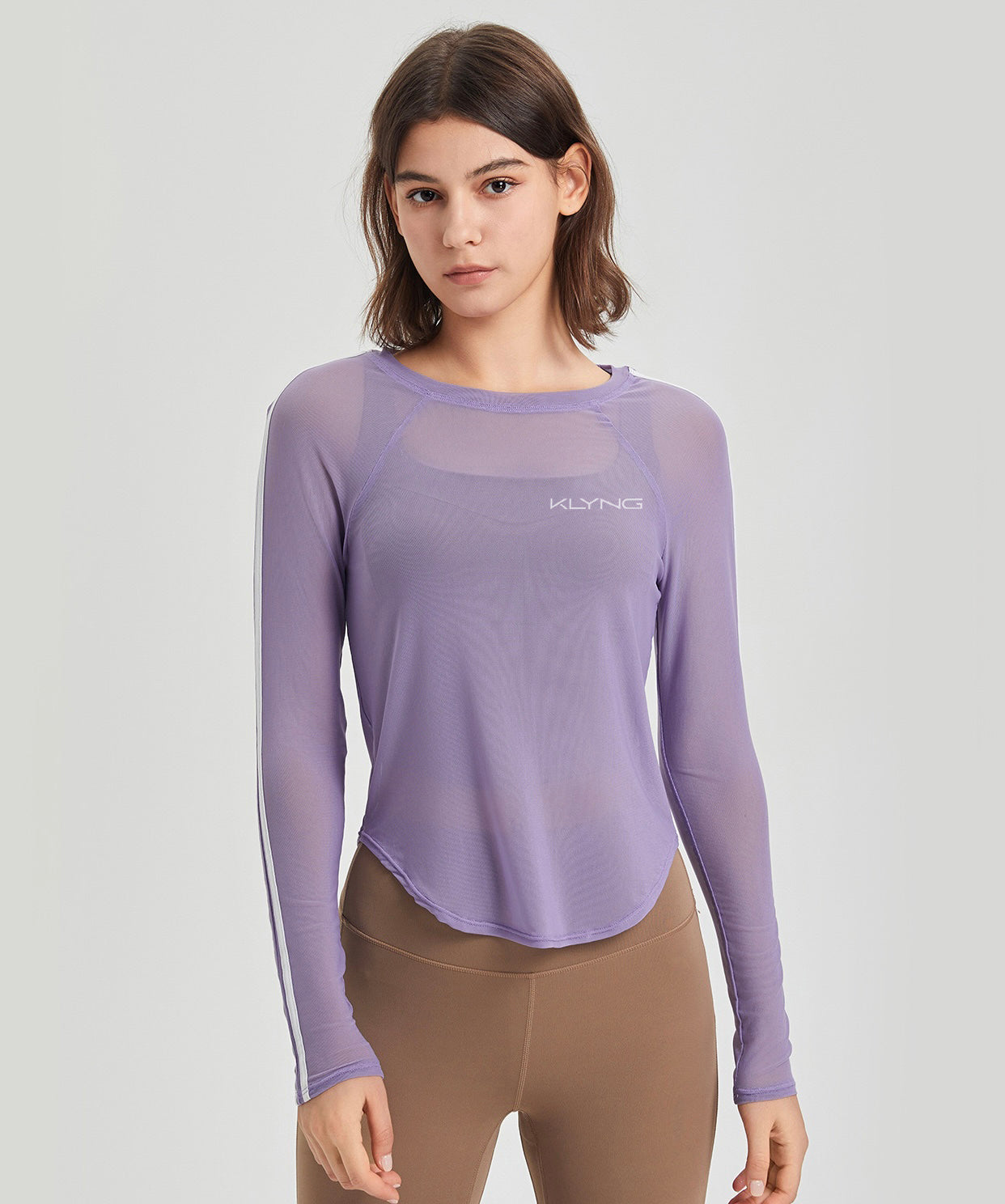 A person wearing the SwiftMesh Long Sleeve Performance Top by Klyng, featuring a sheer lavender fabric and a subtle chest logo, pairs it with brown leggings. The athletic fit is perfect for activities like pickleball against a plain, light-colored background.