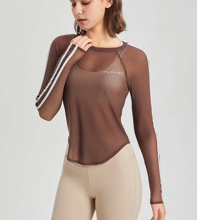 A person dressed in a Klyng SwiftMesh Long Sleeve Performance Top, featuring a brown color with white stripes on the sleeves, and paired with beige leggings. The sheer mesh fabric is adorned with a logo on the chest as they adjust their sleeve and gaze thoughtfully downward.