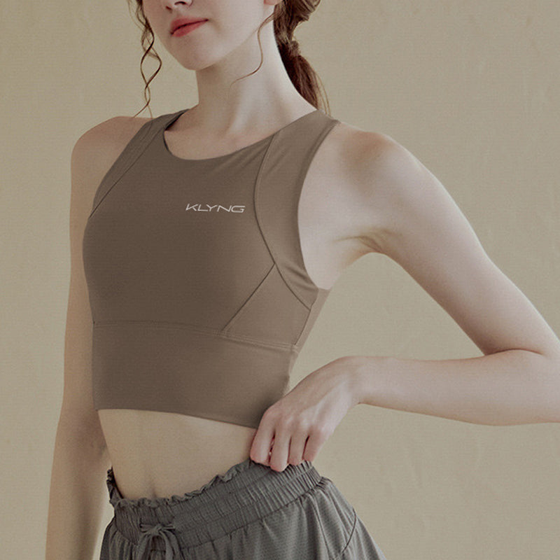 A person is wearing a Women’s PowerFlex Performance Top with a built-in bra and loose gray workout pants, posing with one arm on their hip. The moisture-wicking top, showcasing the brand name Klyng printed on it, ensures comfort while staying active. The backdrop is a plain, light-colored wall.