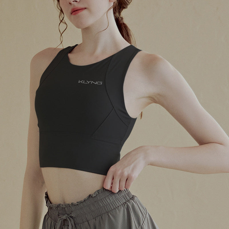 A person wearing a black sleeveless Women’s PowerFlex Performance Top from Klyng, featuring a built-in bra and moisture-wicking technology, pairs it with gray pants. They pose confidently against a beige background, with one hand resting casually on their waist.