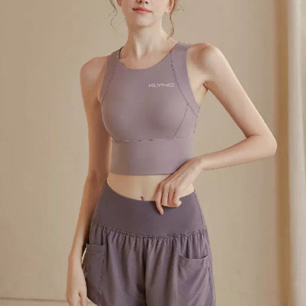 A person standing indoors is dressed in a matching grey Women’s PowerFlex Performance Top with built-in bra from Klyng and shorts set. The moisture-wicking top displays Klyng branding on the chest. The individual has their hands resting on their hips against a neutral-toned background.