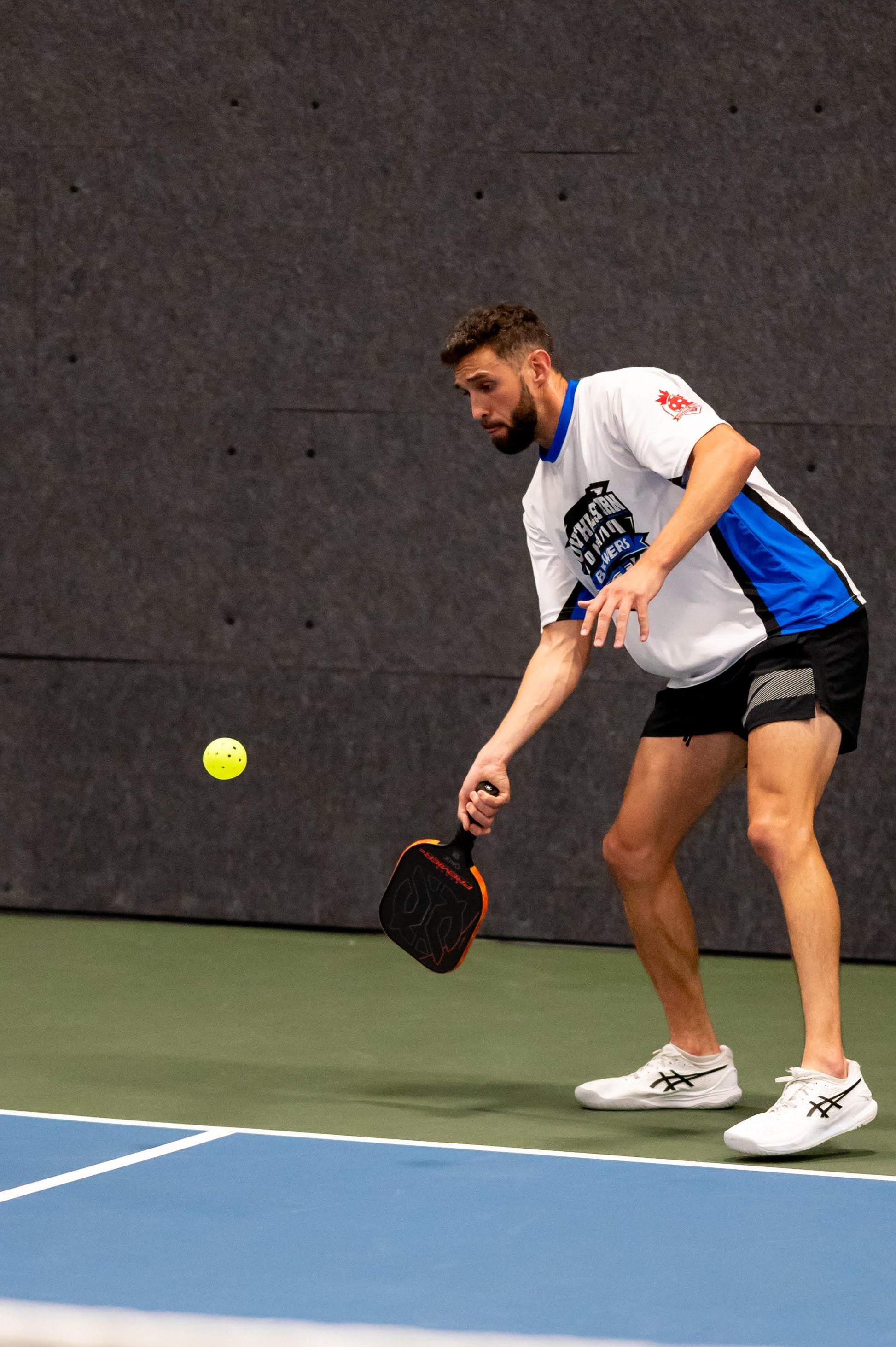 A man wearing a white and blue shirt and black shorts prepares to hit a yellow pickleball with his Evoke Premier Pro Raw Carbon Pickleball Paddle by Onix. He is on an indoor court, focused intently on the ball mid-air.