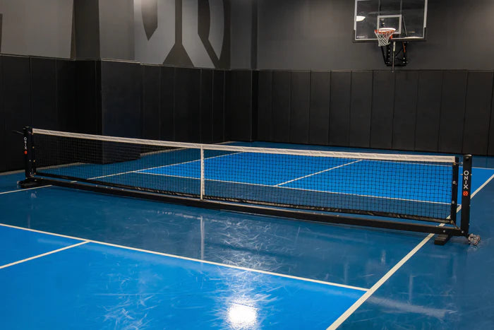 Indoor court featuring a blue floor and the Onix Pro Pickleball Net at the center, suitable for both pickleball and badminton. A basketball hoop is mounted on the wall, with padded panels lining the lower walls. This setup is ideal for both indoor and outdoor use, offering a heavy-duty base for stability.