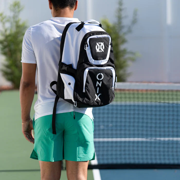 A person in a white shirt and green shorts stands on a tennis court with their back to the camera, carrying an Onix Pro Team Pickleball Backpack in black and white, known for its durability and ample storage. A tennis net and trees are visible in the background.