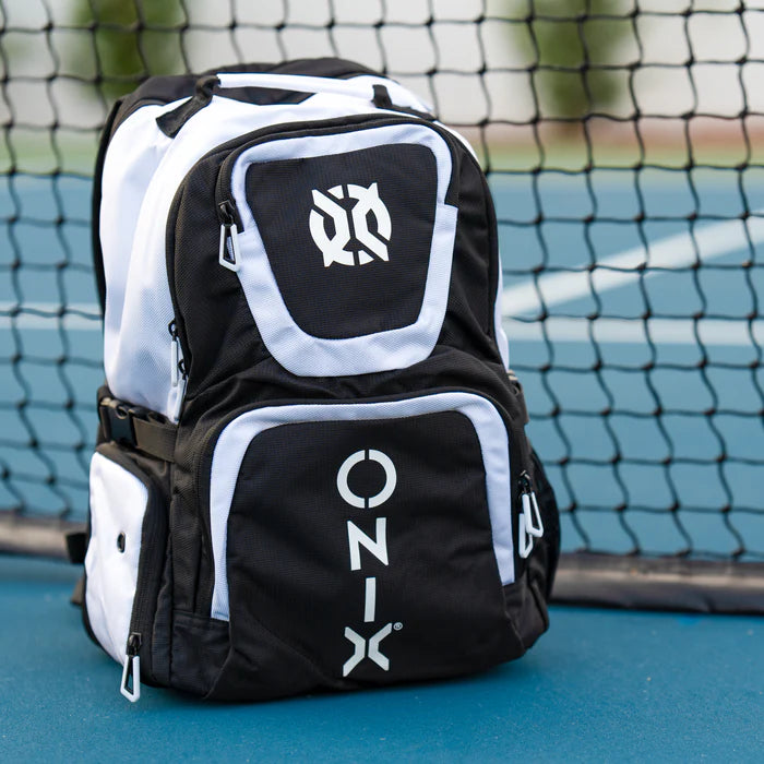 An ONIX Pro Team Pickleball Backpack in black and white stands upright against a tennis net on a blue court. Made from durable polyester, it offers plenty of storage with multiple zippered compartments, featuring the prominent ONIX logo and name on the front.