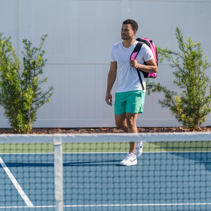 A man in a white t-shirt and green shorts walks on a tennis court, his Pro Team Pickleball Backpack by Onix slung over his shoulder. The bag, known for its ample storage and long-lasting durability, complements the scene with small trees and a white fence in the background.