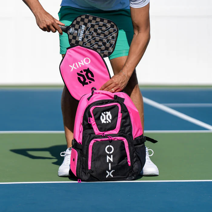 On a blue and green court, a person in green shorts slides a paddle into the Pro Team Pickleball Backpack by Onix, distinguished by its ample storage space and black and pink design. They hold another paddle in their hand.